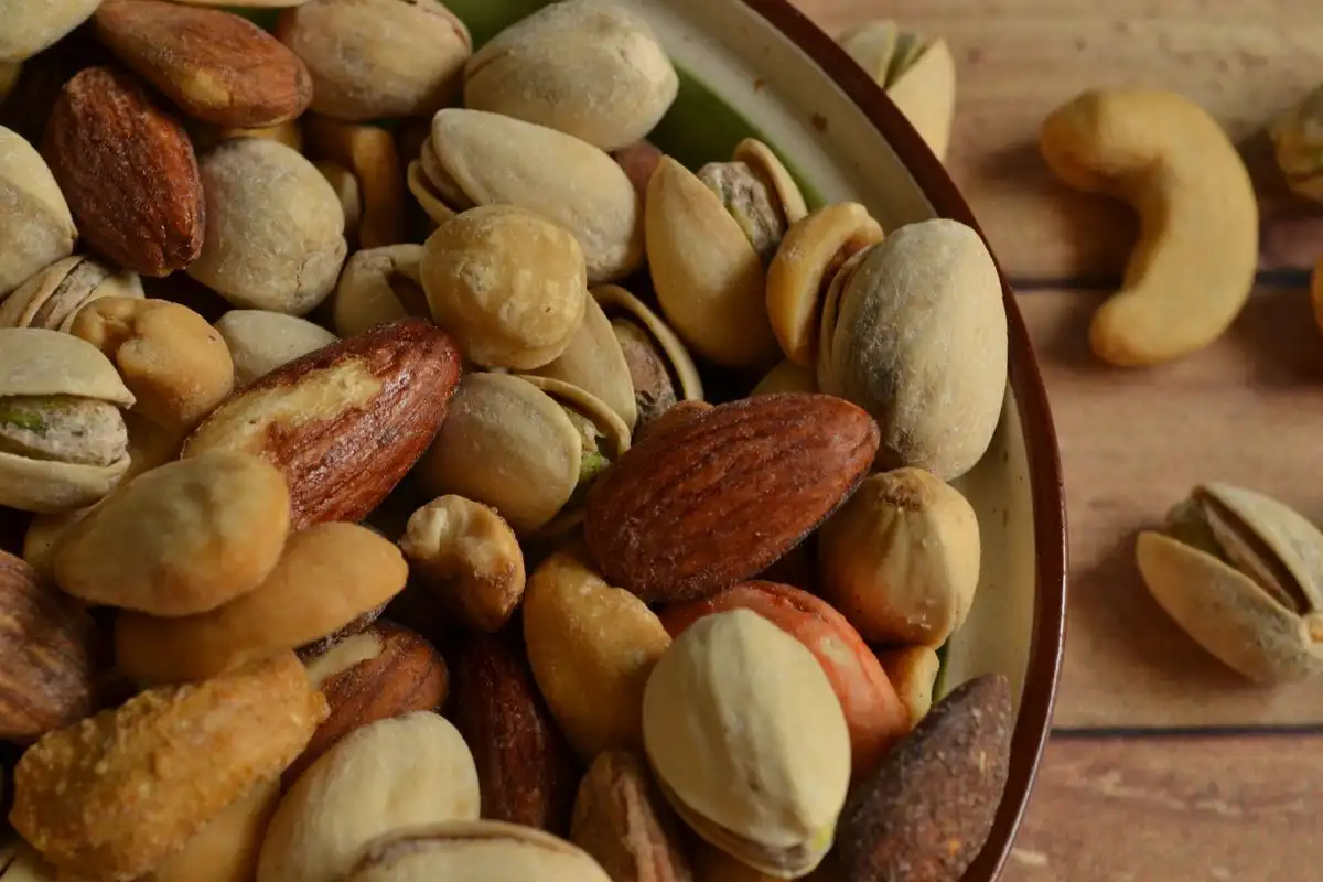 A close-up of a bowl of mixed pistachios in the shell, almonds and cashews on a wooden table