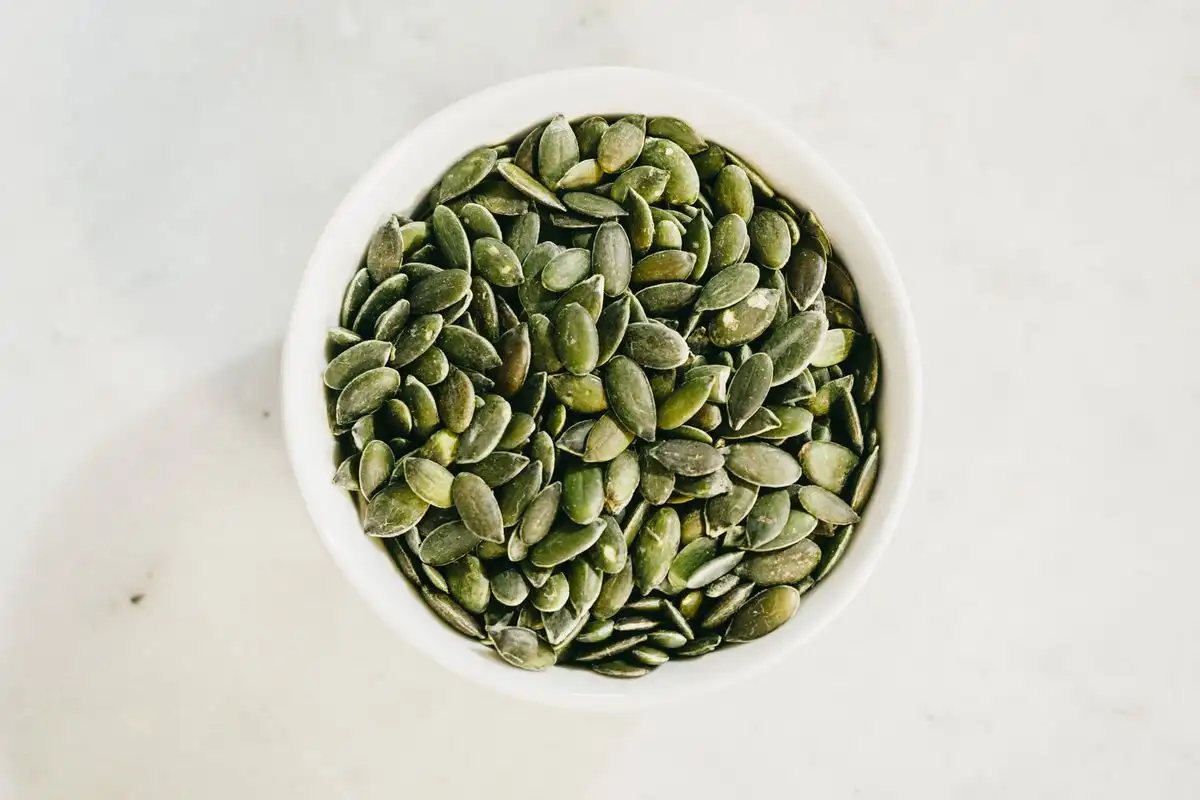 A white bowl filled with green hulled pumpkin seeds on a pale marble surface, seen from above