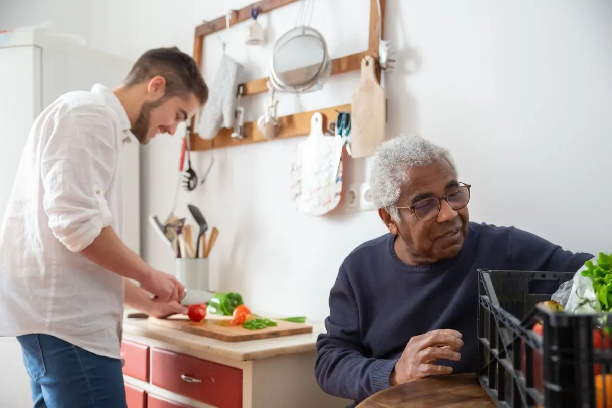 A gray-haired man in glasses unpacking a crate of fresh vegetables in a home kitchen while a younger man slices tomatoes and peppers