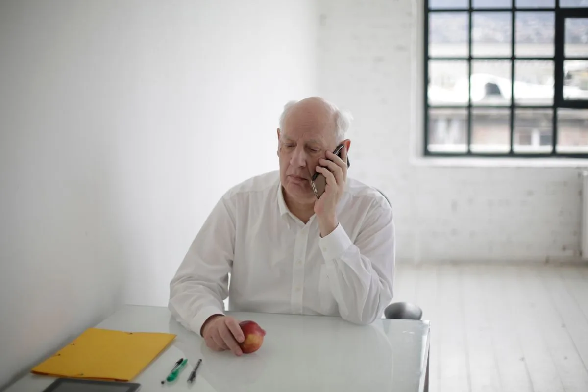 A white-haired man in a white shirt talking on his phone at a bright table, a yellow folder and pens beside him