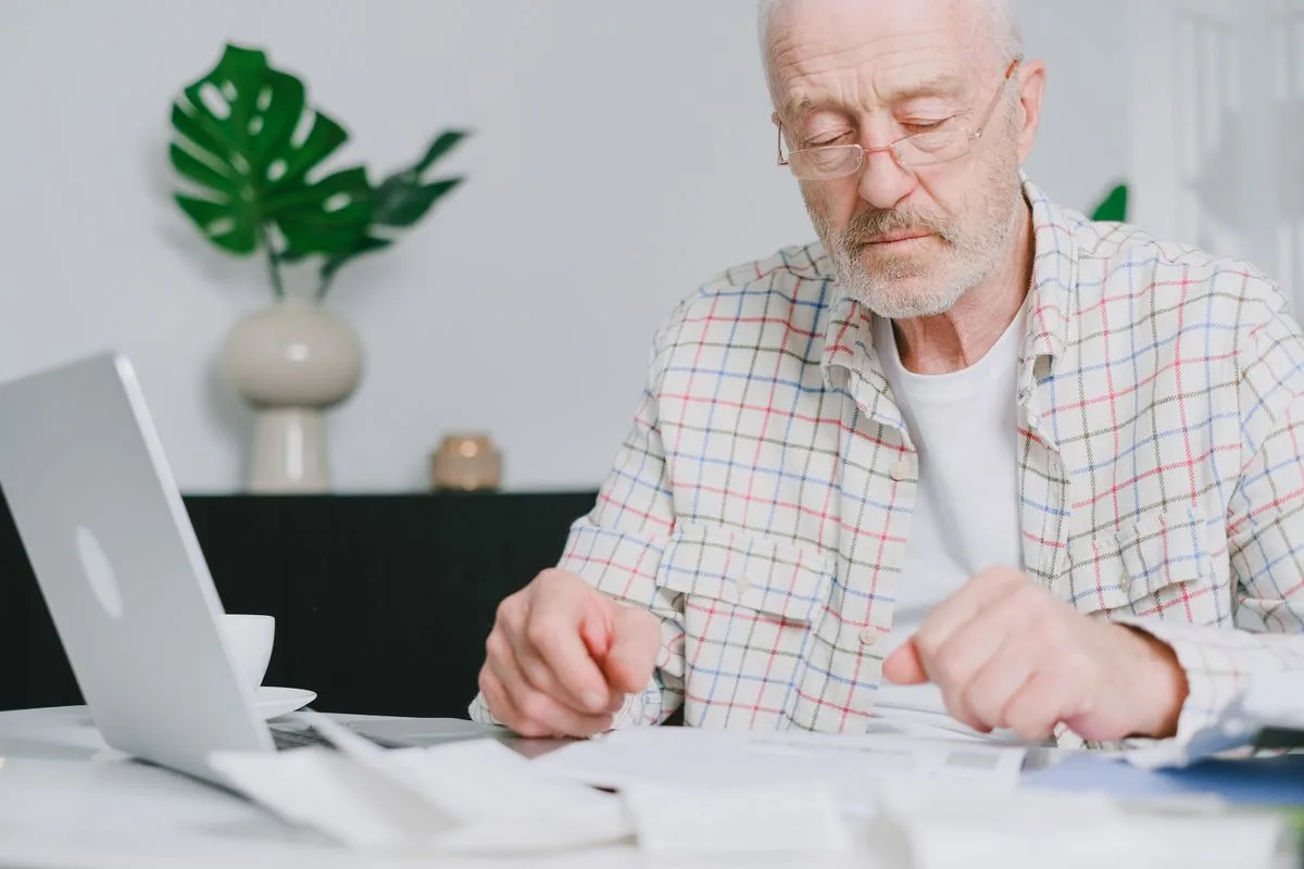 A white-haired man in his sixties with glasses carefully reading printed papers at a table beside a laptop