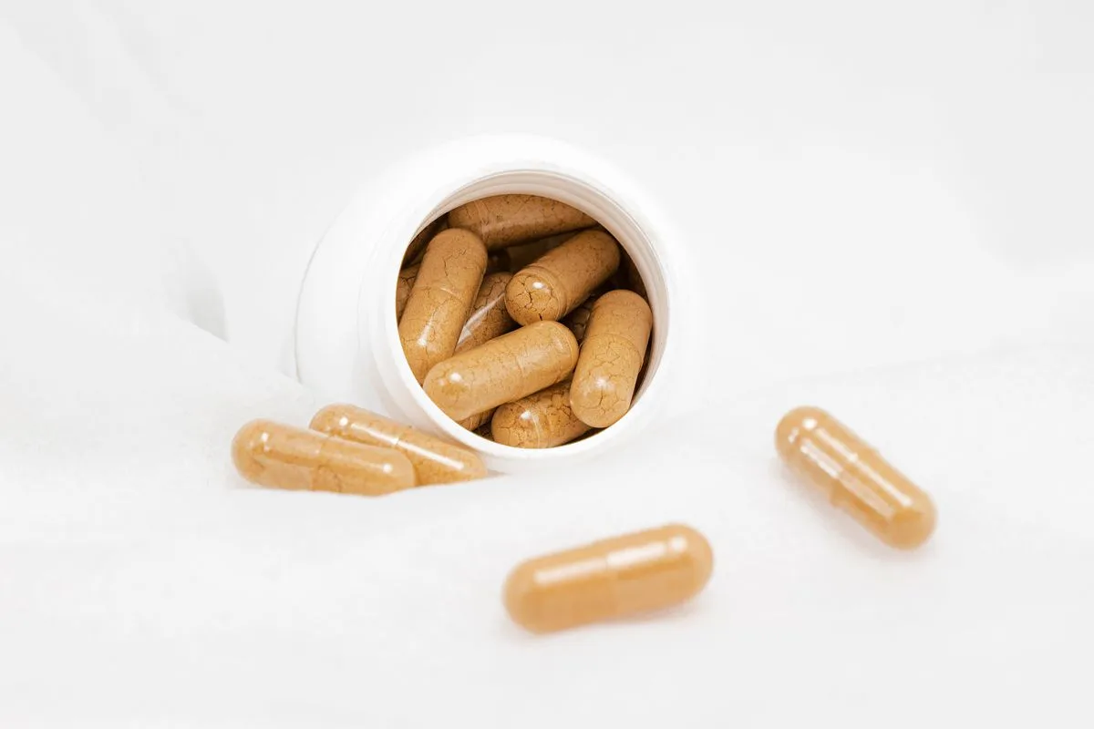 Tan herbal capsules spilling out of a small white supplement bottle lying on a white cloth