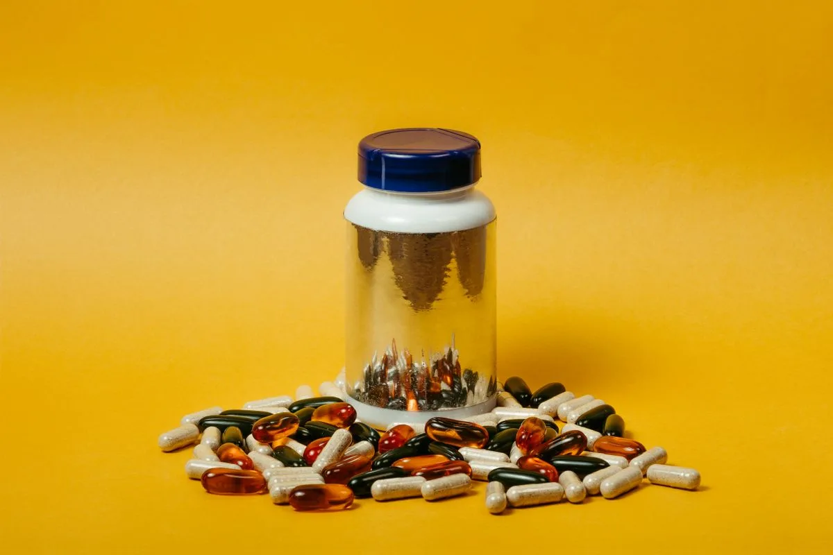 A clear supplement bottle filled with capsules standing on a bright yellow background, surrounded by spilled amber softgels and white capsules