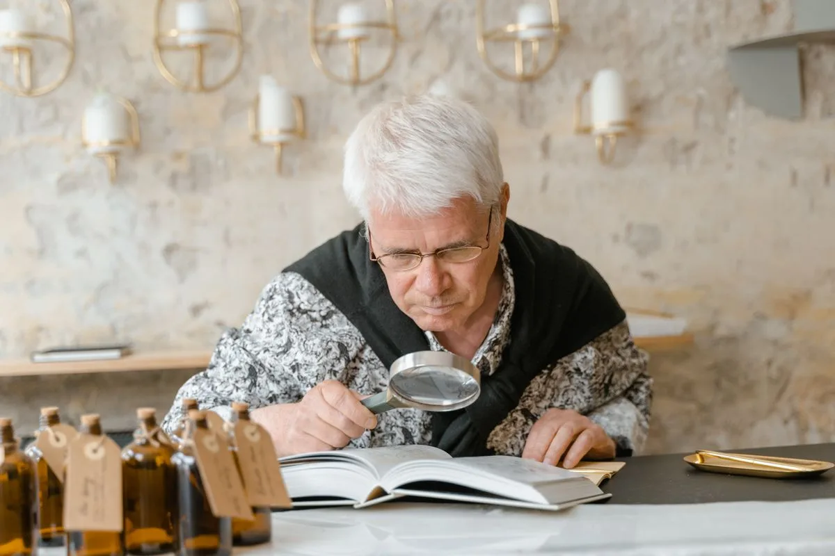 A white-haired man in glasses leaning over an open book with a magnifying glass, a row of amber bottles on the table in front of him