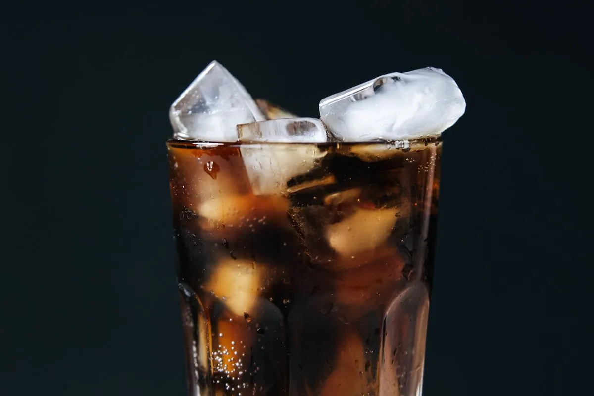 A tall glass filled with dark cola and ice cubes photographed against a deep navy background