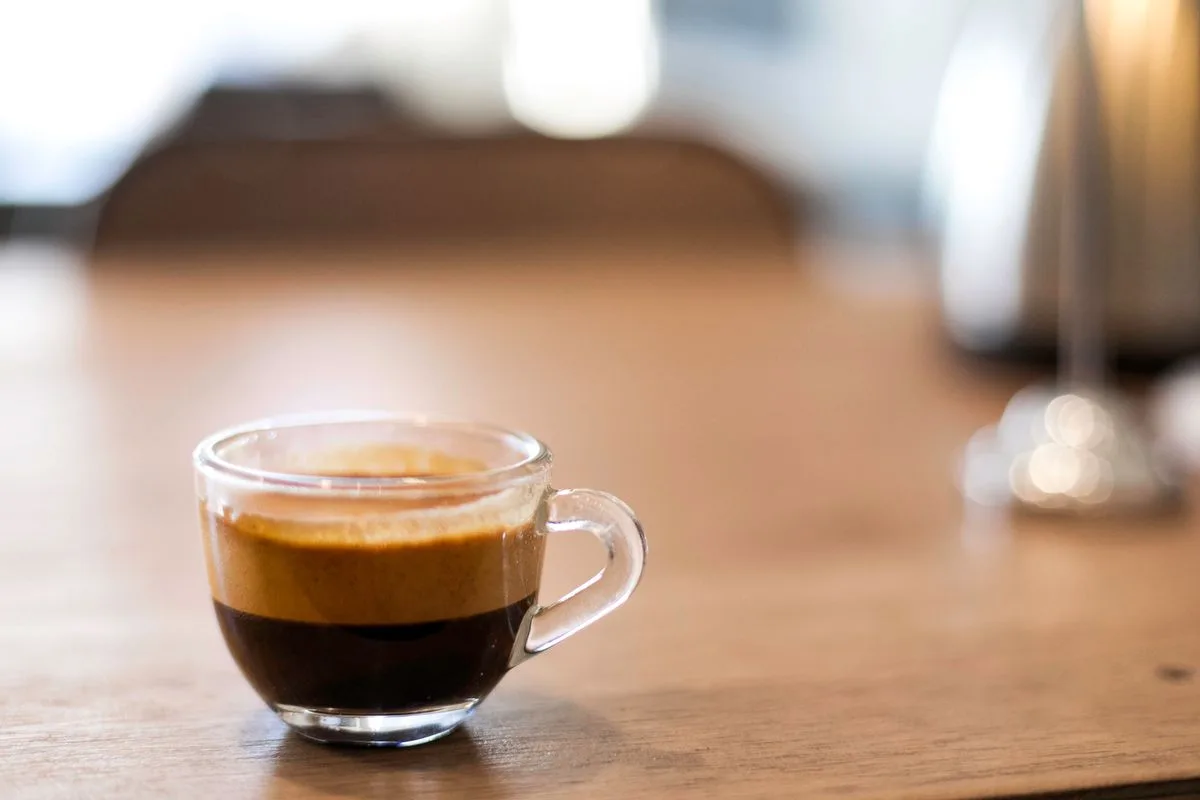 A small glass cup of freshly brewed espresso resting on a dark wooden table in low warm light