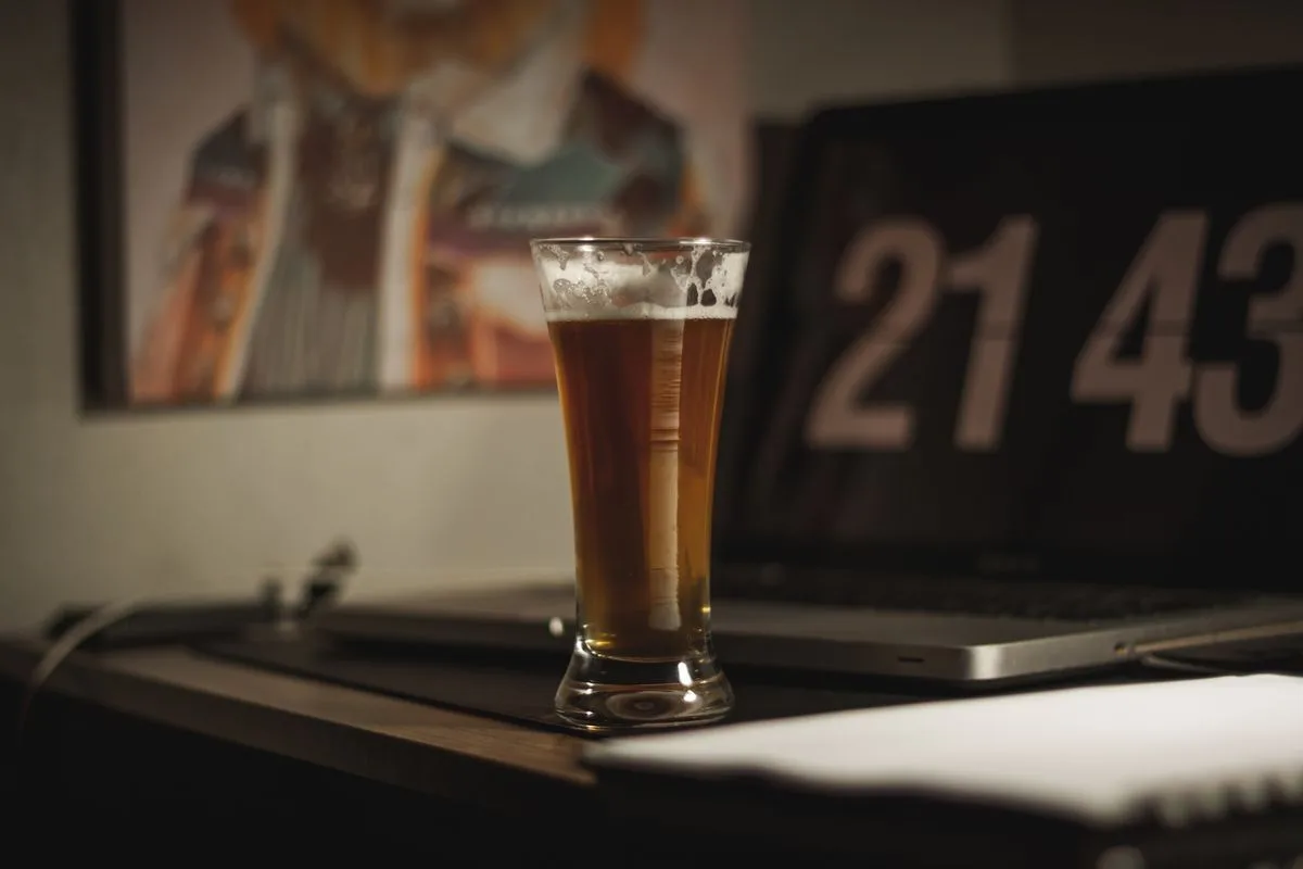 A tall glass of beer standing on a dark wooden desk in a dim room, with a screen behind it showing the time as 21:43