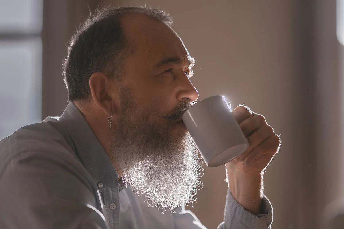 A man in his mid-fifties with a grey beard drinking from a mug in warm morning light, looking thoughtfully out of the window