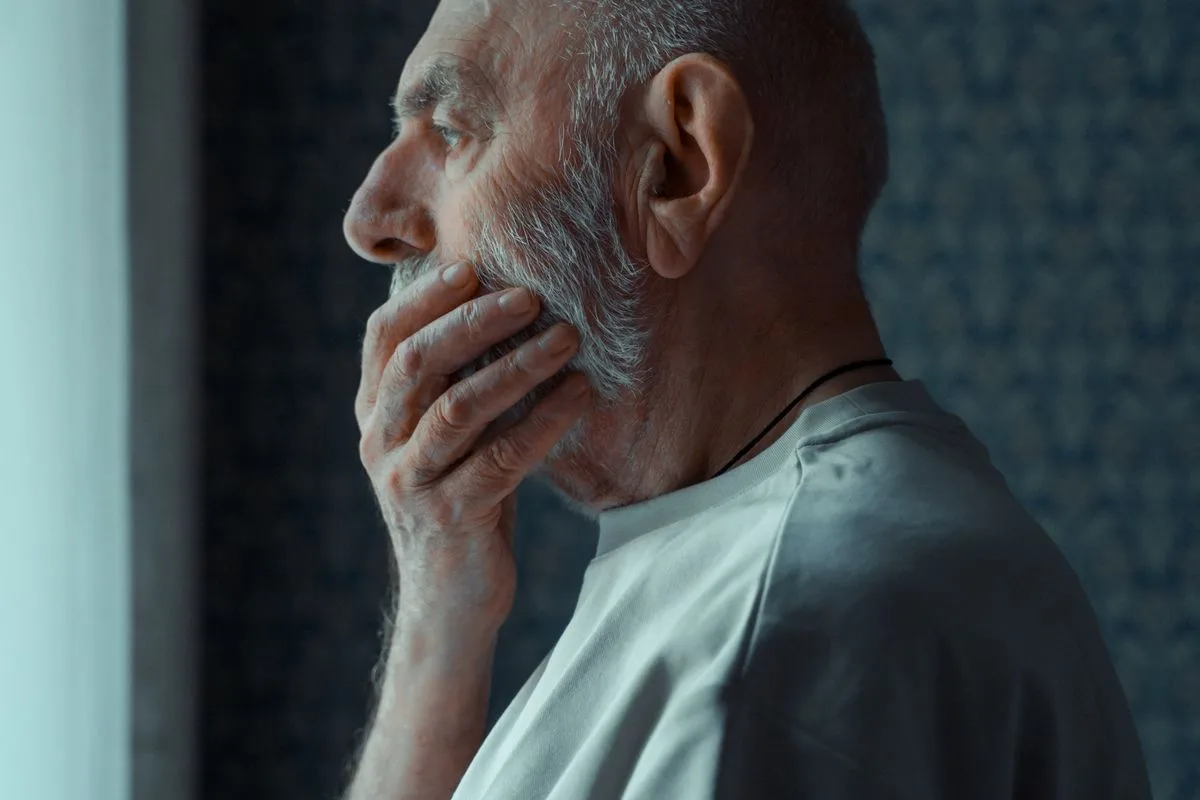 An older man with a white beard standing by a window at home, hand on his chin, looking away thoughtfully