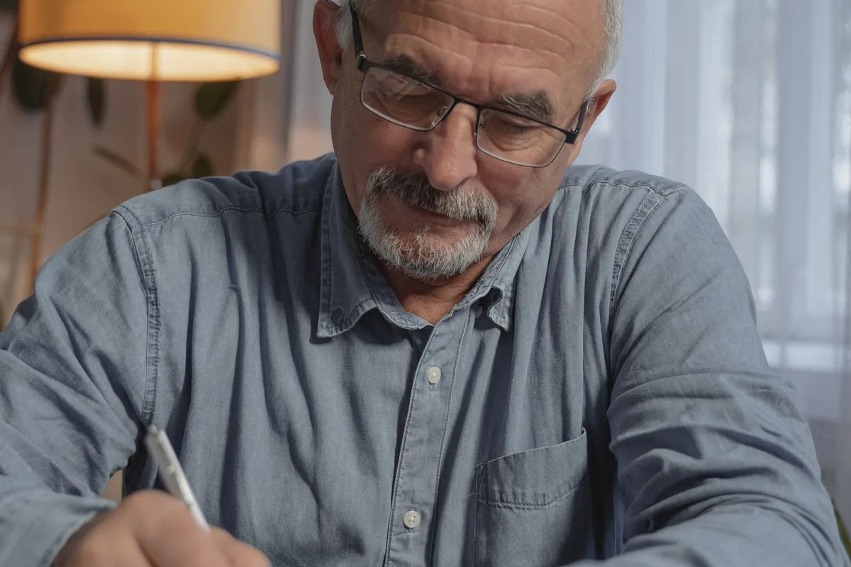 An older man with grey hair, glasses and a denim shirt writing in a notebook at a table by a lamp at home