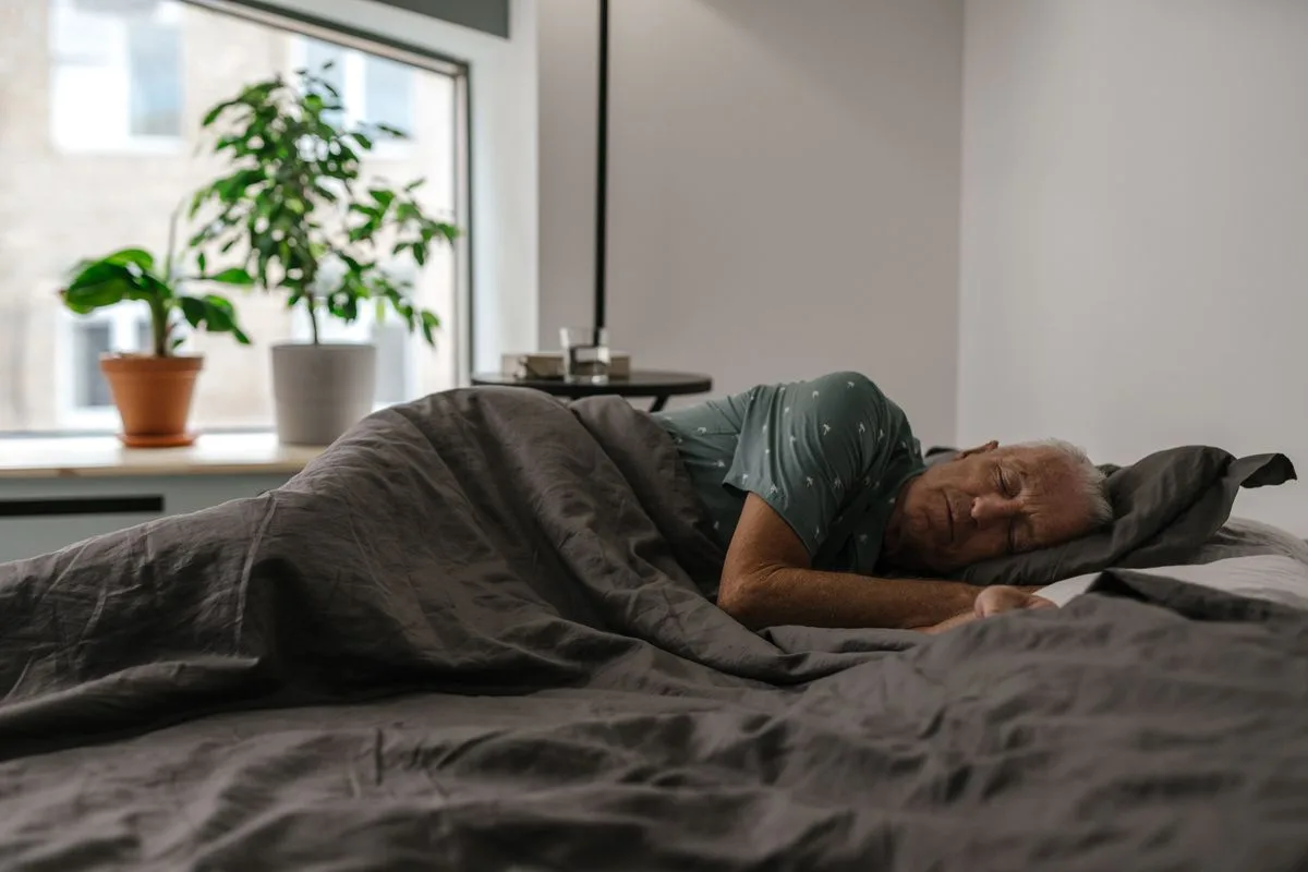 An older man with grey hair sleeping peacefully on his side in bed in a softly lit bedroom