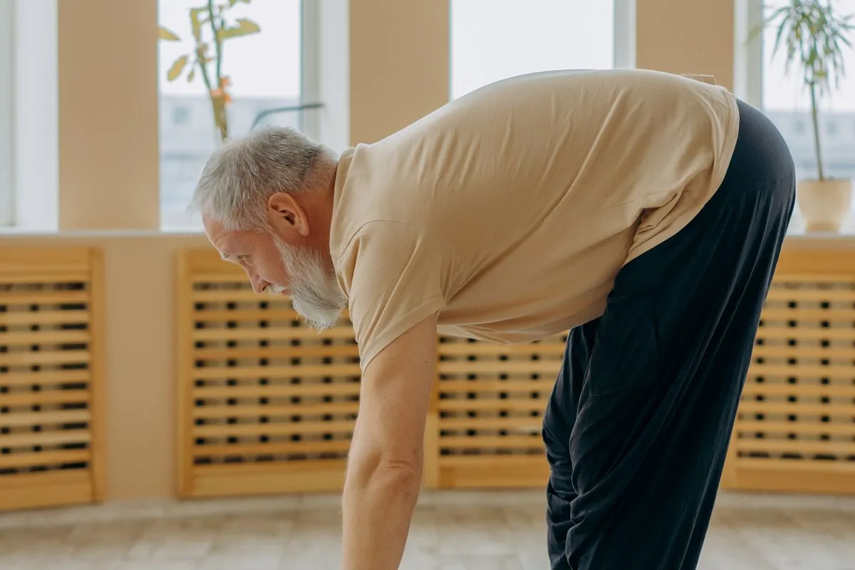 A grey-bearded man in his sixties bending forward during an exercise session in a bright room with tall windows