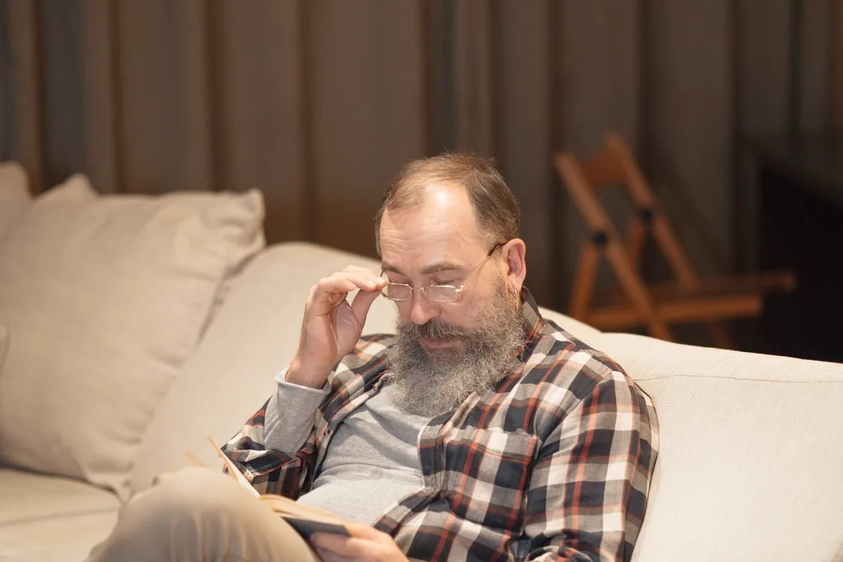 A bearded man in his late fifties sitting on a sofa with a book, adjusting his glasses in a warmly lit living room