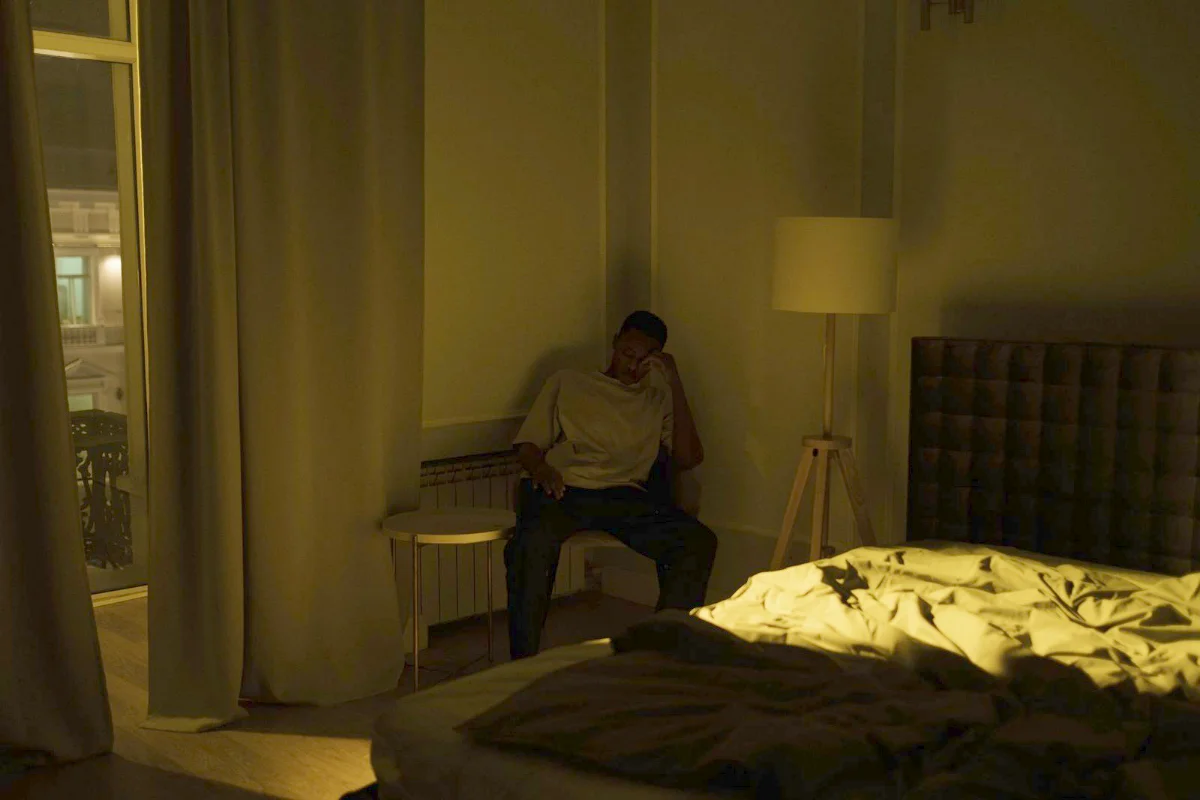 A man sitting awake at the edge of a chair in a dim bedroom at night, head in his hand, with the bed visible in the foreground