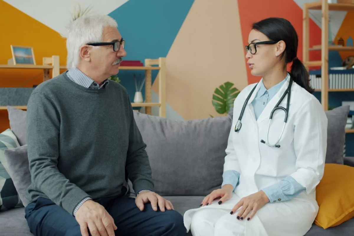 An older man in glasses talking with a female doctor holding a stethoscope during a home consultation