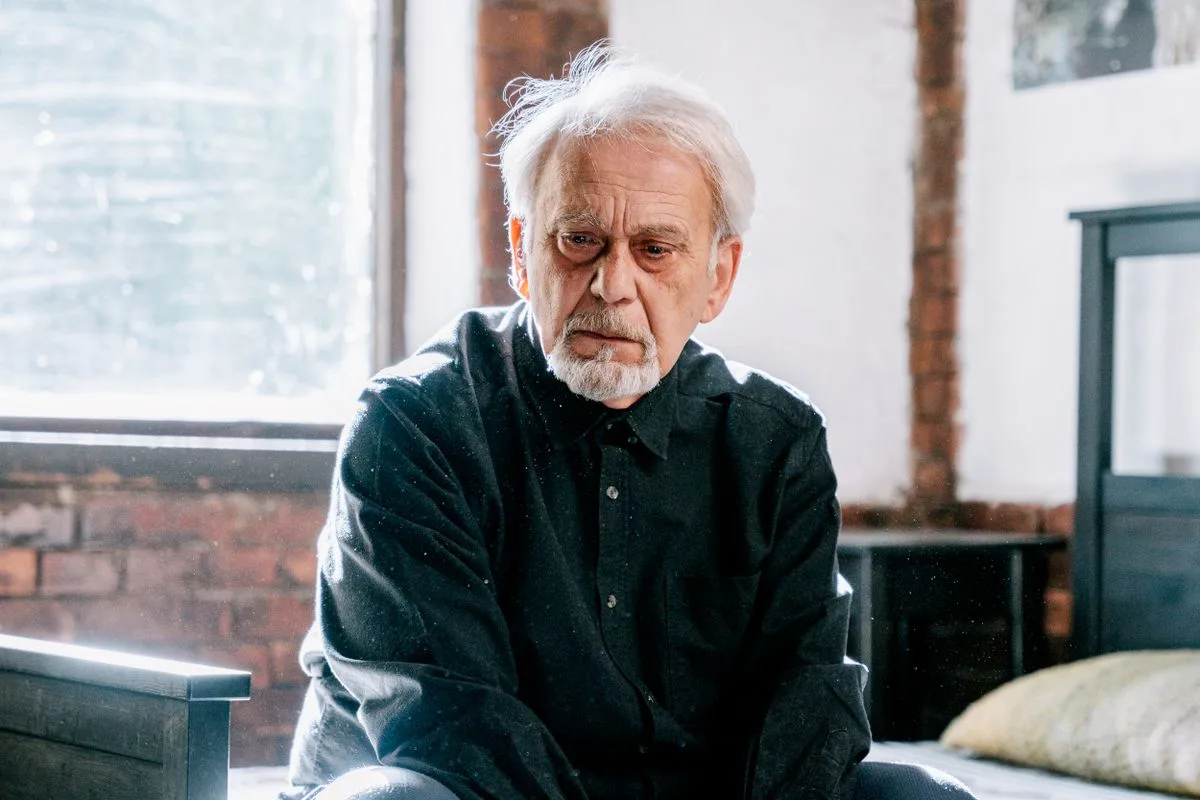 A white-haired man in a dark shirt sitting on the edge of his bed beside a window, looking down thoughtfully