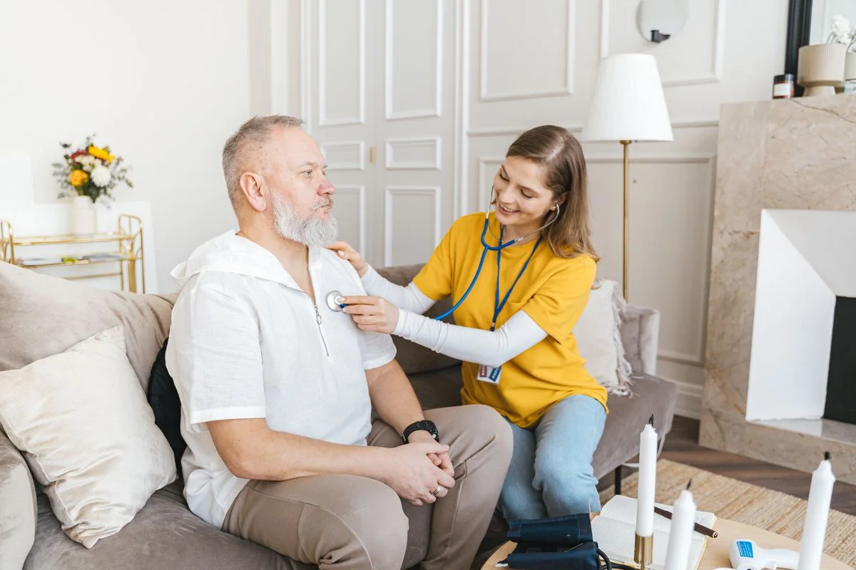 A home health worker using a stethoscope on an older man with white hair and a white beard, seated on a sofa in his living room