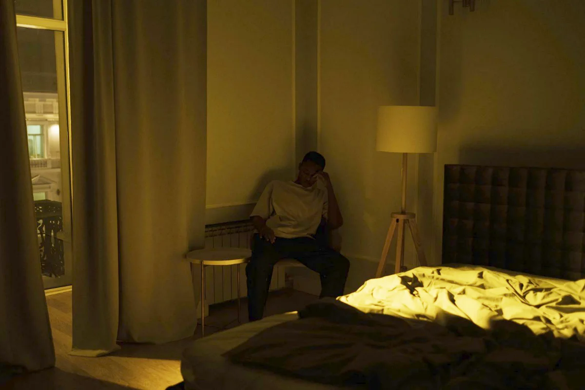 A man sitting awake in a dark bedroom late at night with a lamp on and the bed left unmade behind him