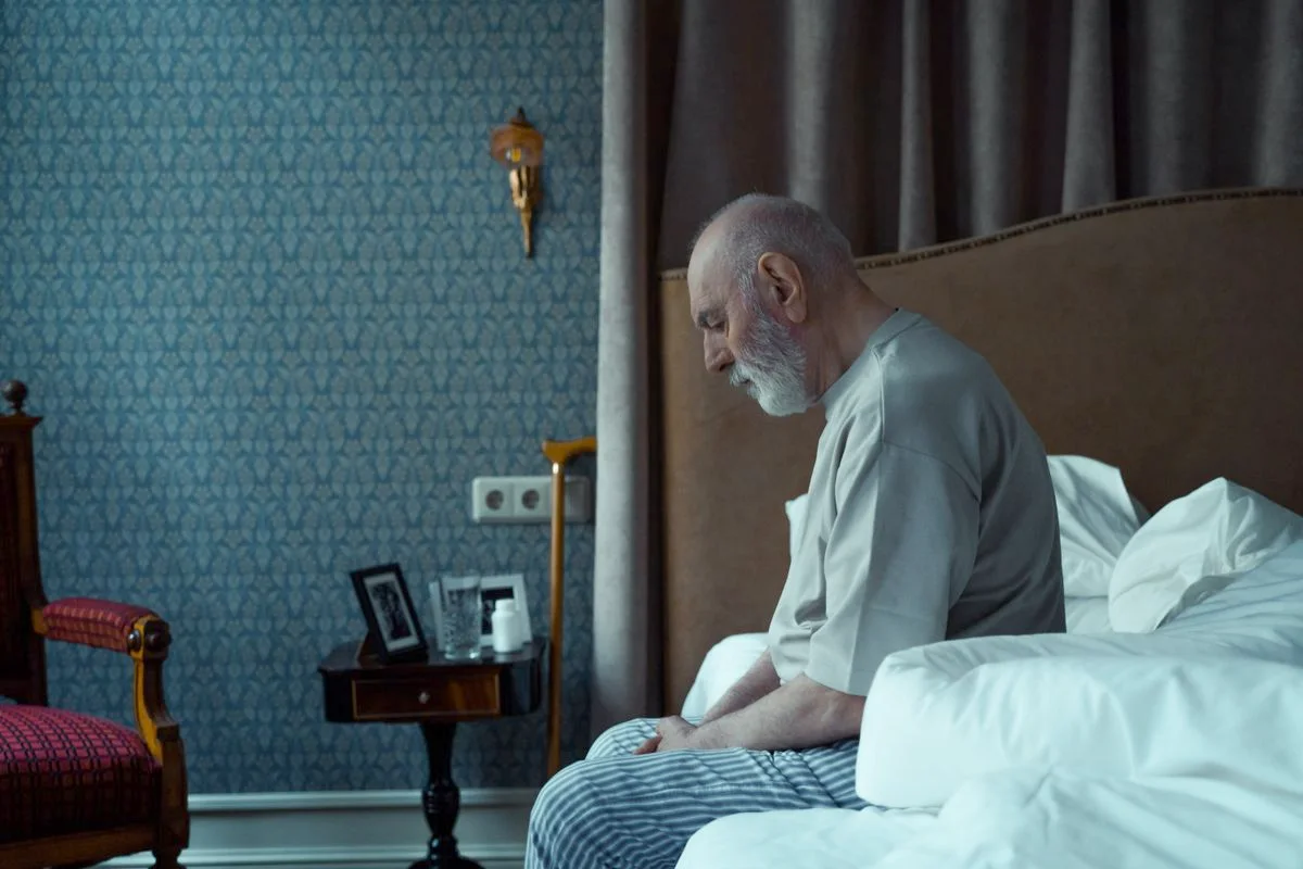 An older man with white hair sitting alone on the edge of his bed at night, head lowered
