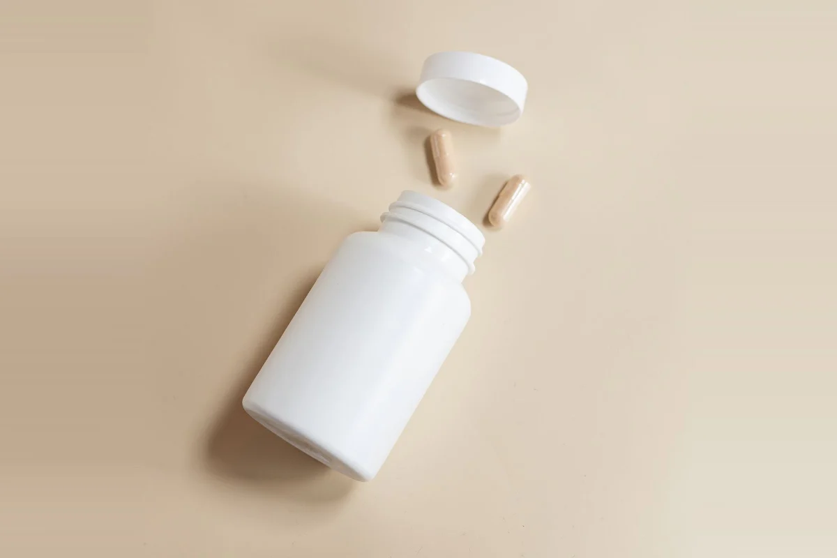 A white supplement bottle on its side with two tan capsules spilled out and the lid removed, on a warm beige background