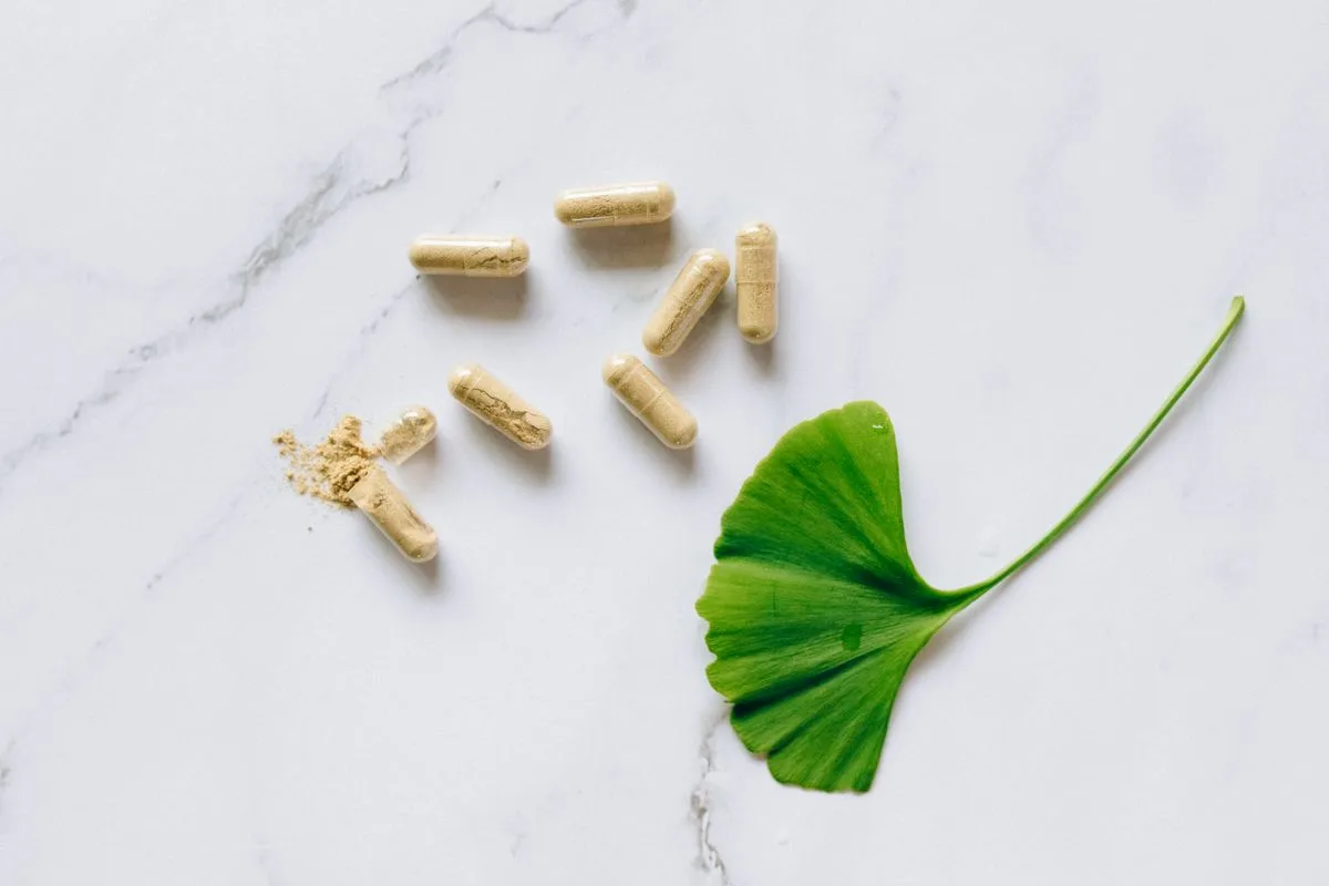 Tan supplement capsules scattered next to a single green ginkgo leaf on a white marble surface