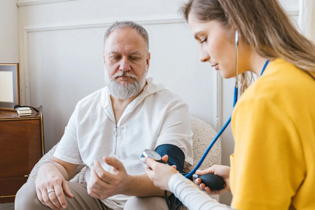 A grey-bearded man in his sixties sitting in an armchair while a clinician in a yellow top takes his blood pressure