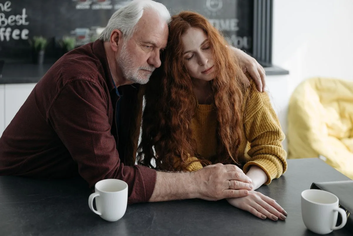 An older man with white hair and a beard sitting at a kitchen table with his arm around an adult daughter, two coffee mugs in front of them