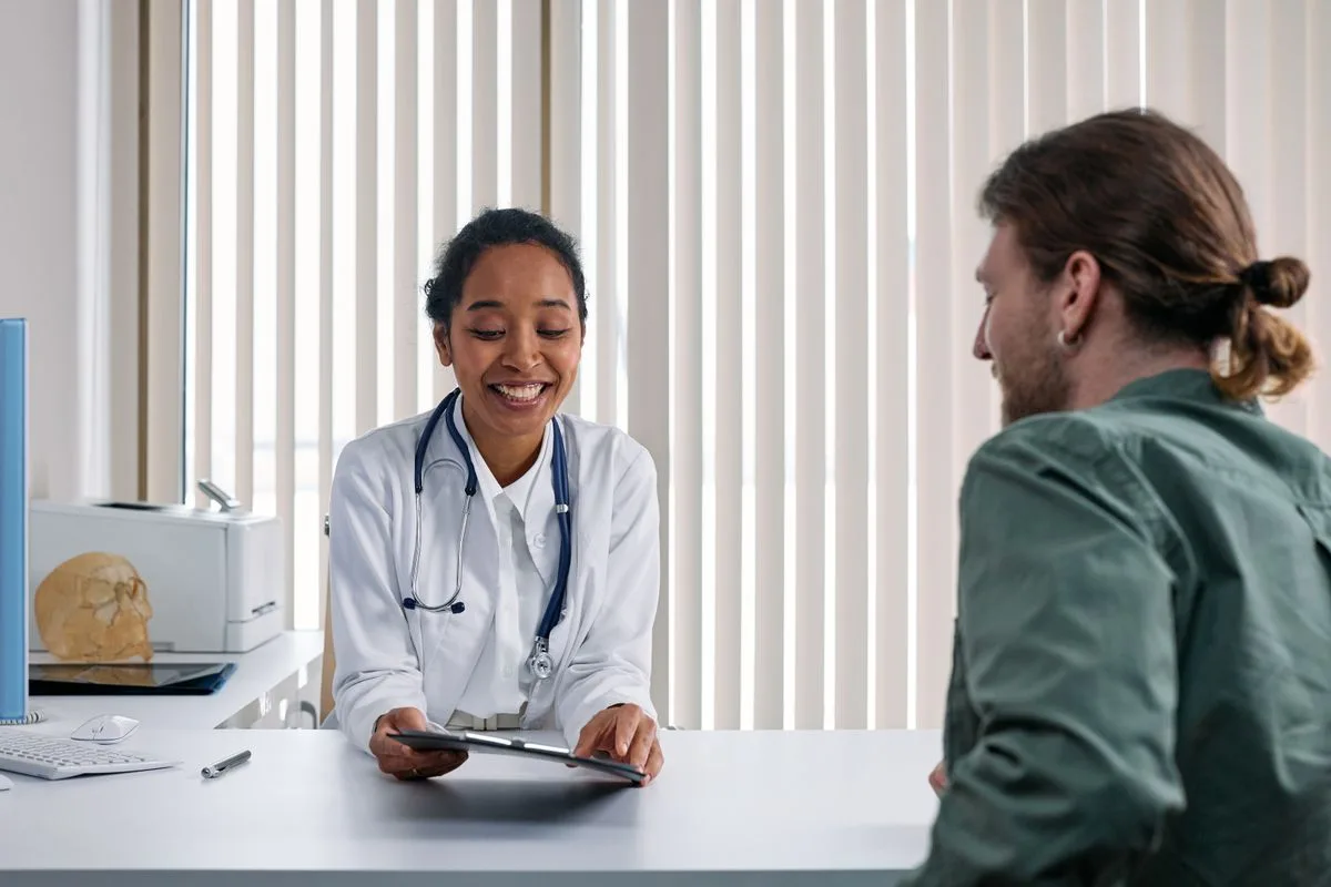 A doctor in a white coat sitting at a desk with a tablet, smiling as she talks with a male patient in a bright clinic office