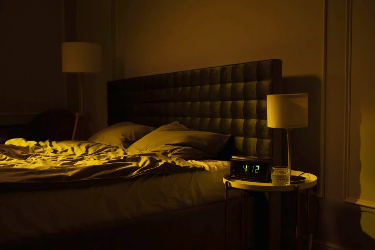 Dark bedroom with an empty bed and a bedside alarm clock reading 4:12 beside a glass of water
