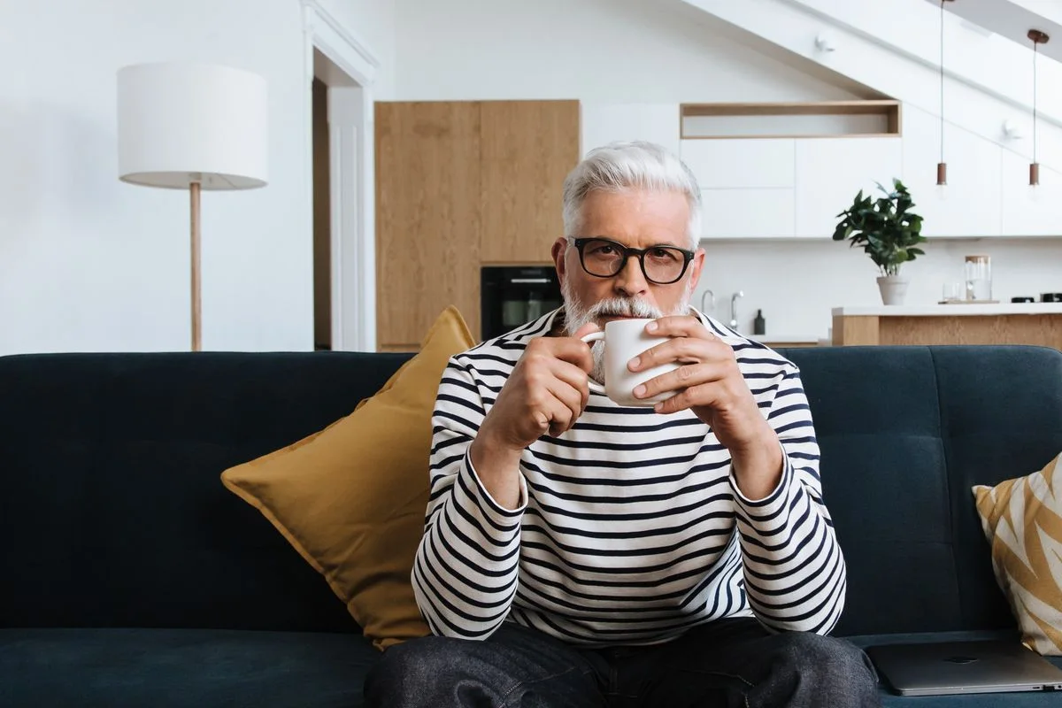 A well-groomed man around fifty with grey hair, glasses and a striped shirt, sitting on a sofa at home with a mug of coffee