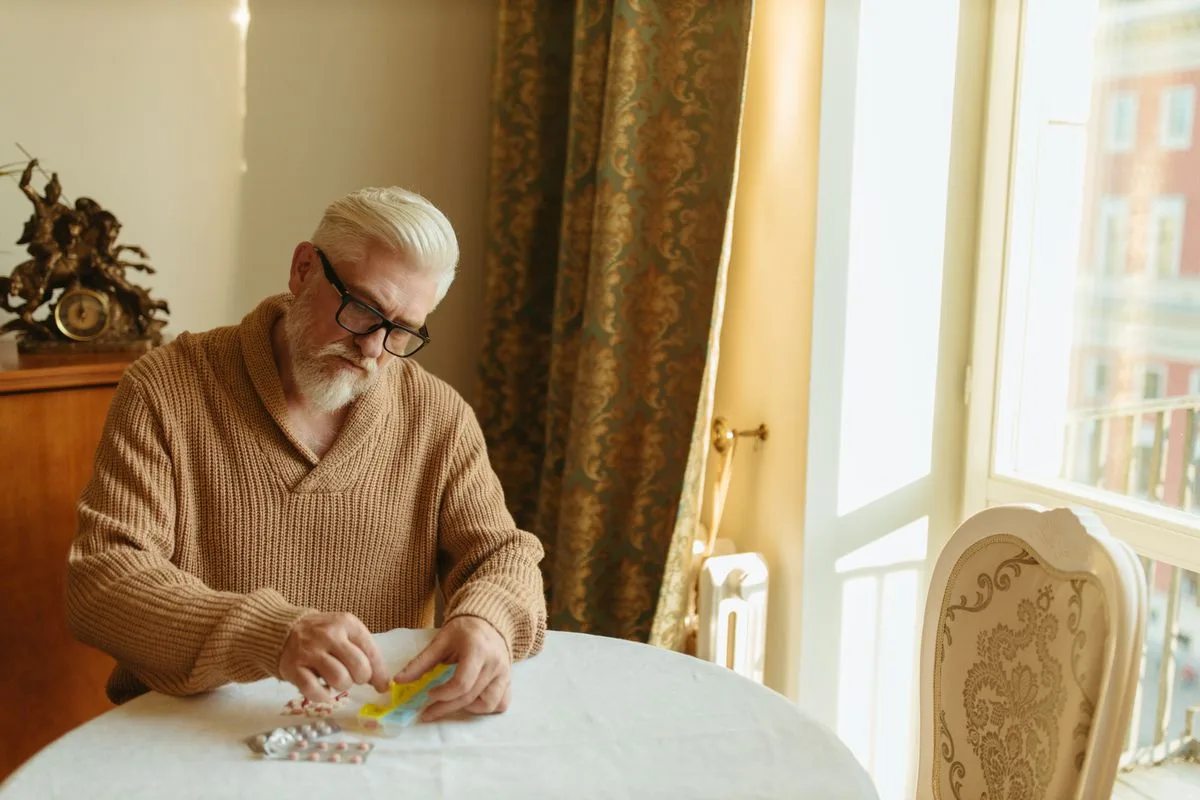 A white-haired man in his sixties wearing glasses, sorting his daily tablets into a weekly pill organiser at a table by a window