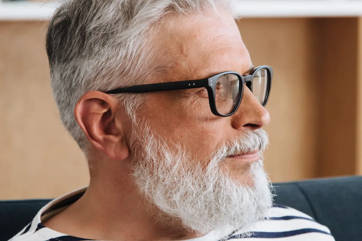 A man in his sixties with grey hair, a full white beard and dark-framed glasses, looking off to the side with a serious expression