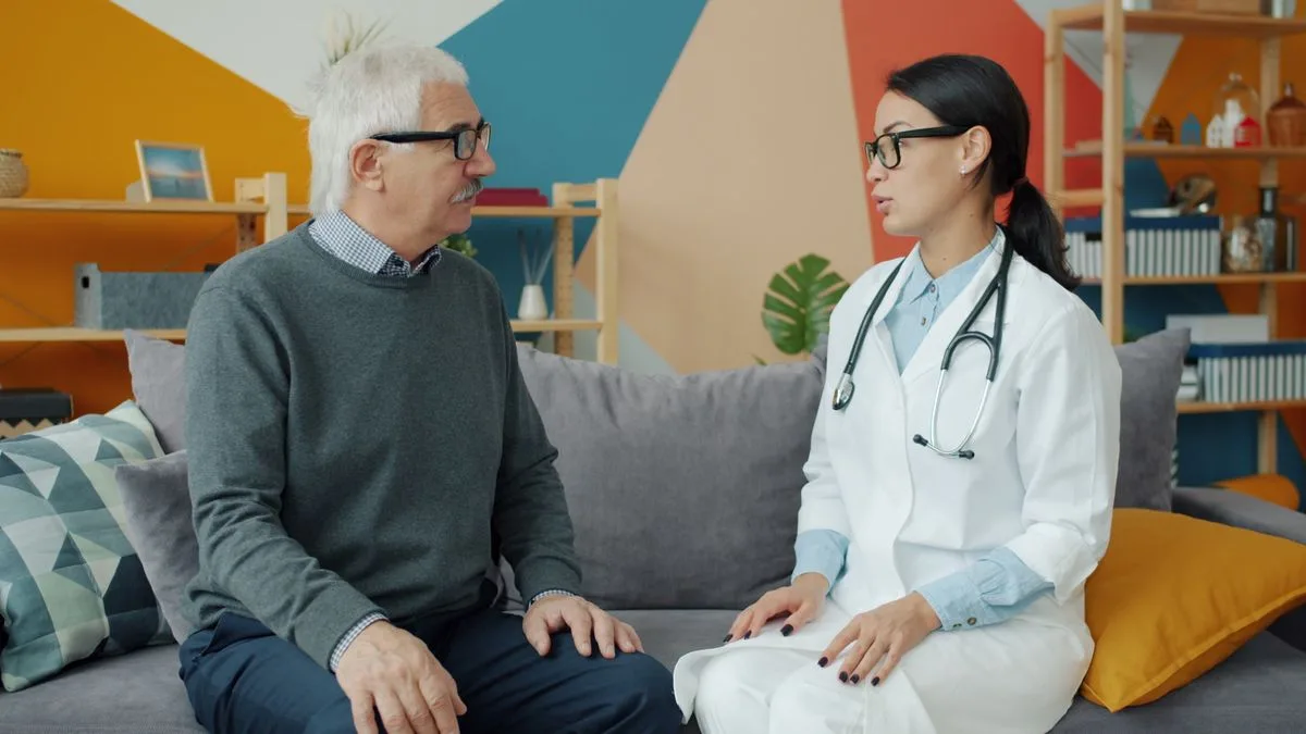 An older man with grey hair and glasses sitting on a sofa talking with a doctor in a white coat with a stethoscope